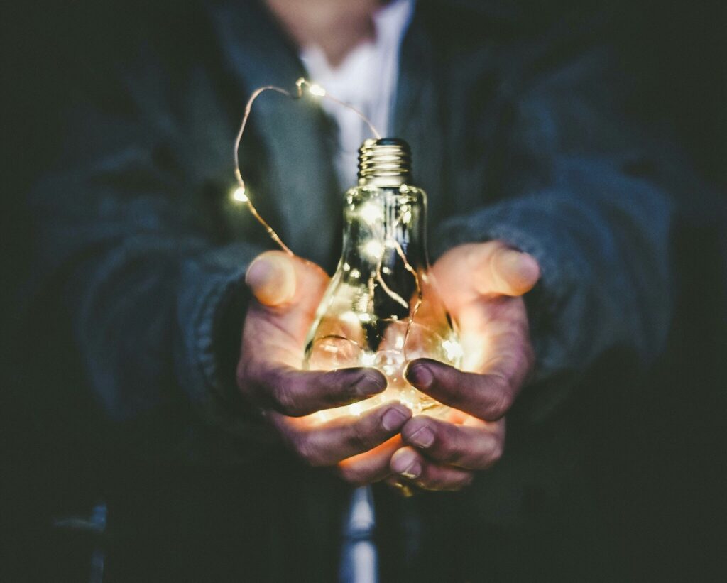 man holding an illuminated lightbulb amongst trees in Stellenbosch, South Africa by Riccardo Annandale (@pavement_special)