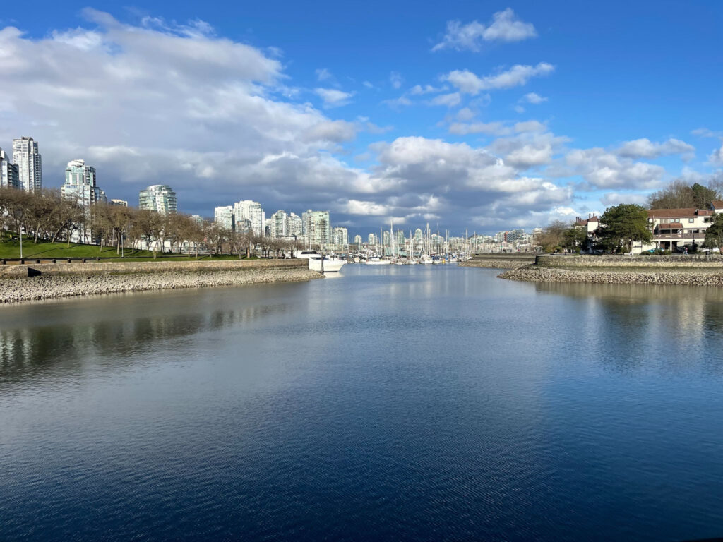 A calm waterfront scene with buildings on the left, trees and grassy areas, and sailboats in a marina on the right, under a partly cloudy sky.